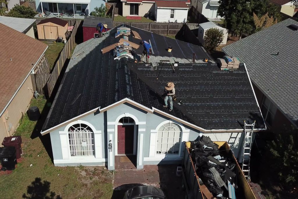 Aerial view of a crew mid re-roof on a single-story home, with old shingles piled in a dumpster in the driveway