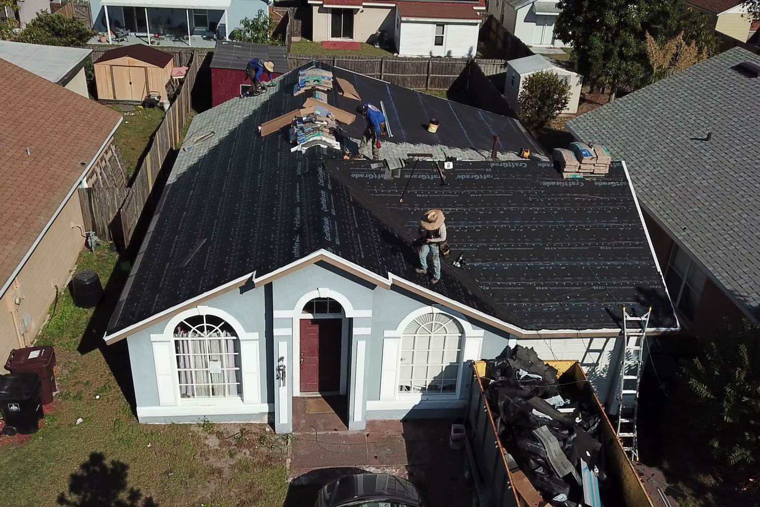 Aerial view of a crew mid re-roof on a single-story home, with old shingles piled in a dumpster in the driveway